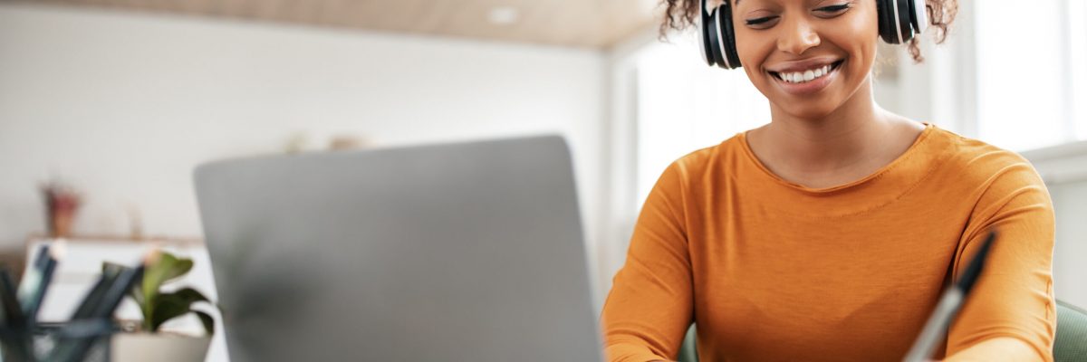 Smiling young black woman in wireless headphones sitting at desk table working on laptop and writing letter in paper notebook, taking notes watching weninar, holding pen in hand, free copy space